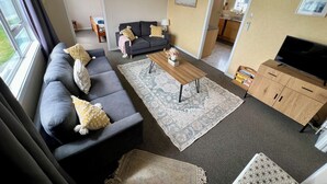 Un salon avec un canapé, une table basse et un téléviseur. On aperçoit un coin cuisine à travers une porte ouverte.