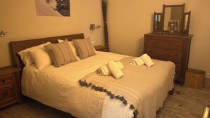 Una cama bien tendida con un edredón blanco y varias almohadas, un aparador de madera con un espejo y un cuadro colgado en la pared de un paisaje nevado.