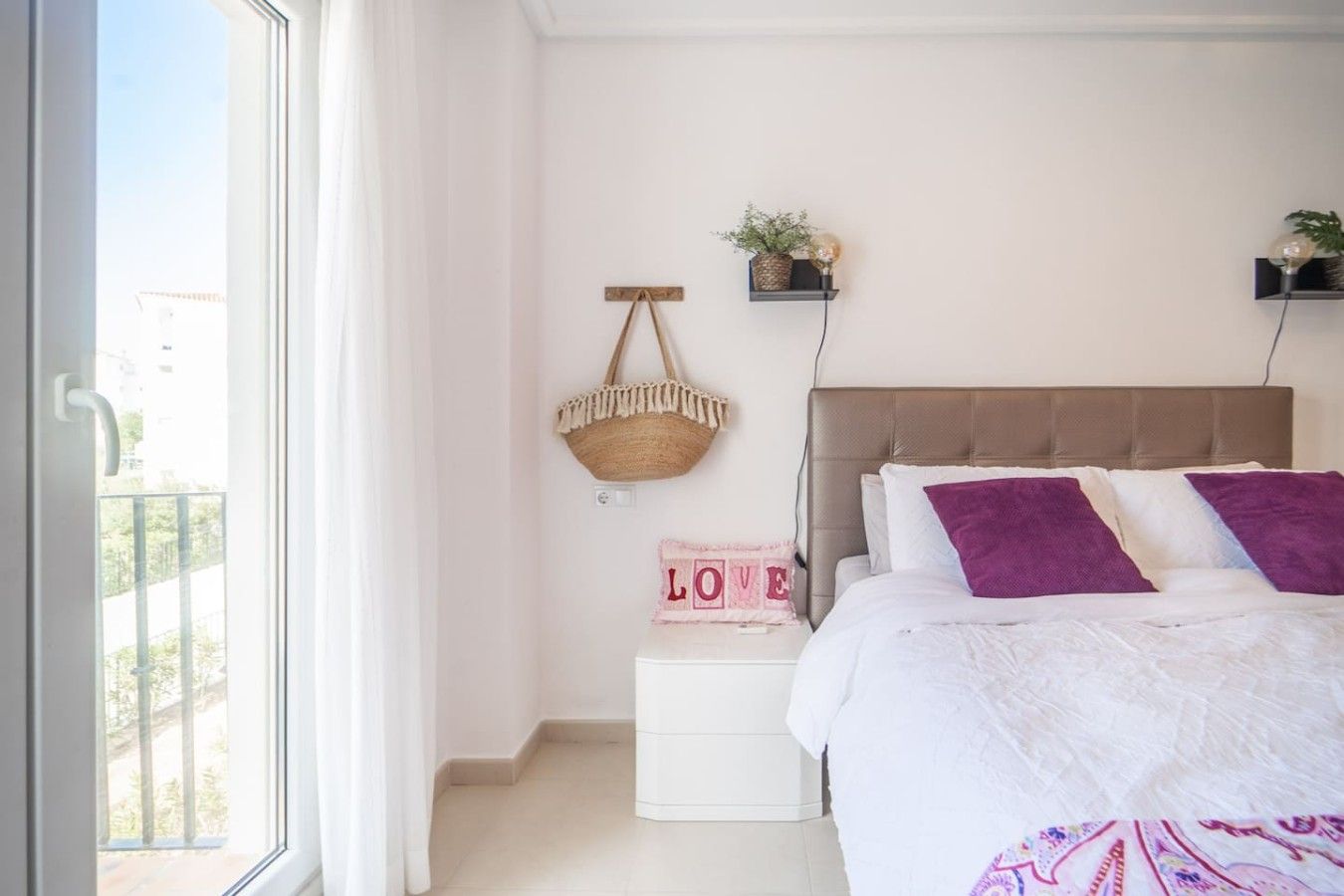A bedroom with a bed, a bedside table, a hanging basket, and a wall-mounted shelf.
