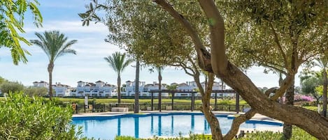 A swimming pool surrounded by lush greenery and palm trees, with a row of white buildings in the background.