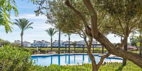 A swimming pool surrounded by lush greenery and palm trees, with a row of white buildings in the background.