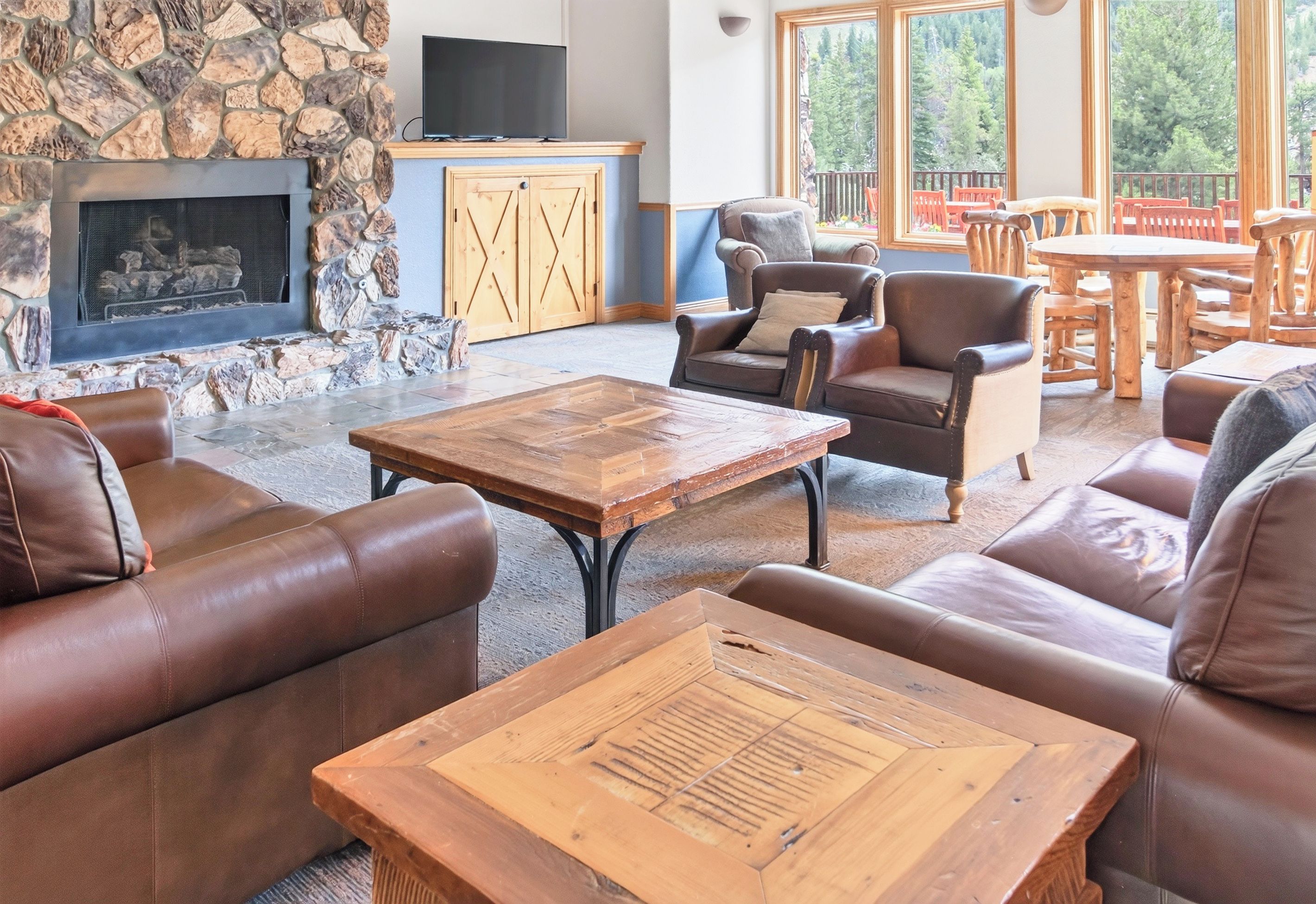 A cozy living room with leather sofas, a stone fireplace, wooden furniture, and a large window offering a view of trees.
