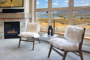 A room with a fireplace, two white furry chairs, a TV mounted on the wall, and large windows with a view of a lake and mountains. - Modern Condo. Hot tub. Amazing Lake views. Mural. (Frisco)