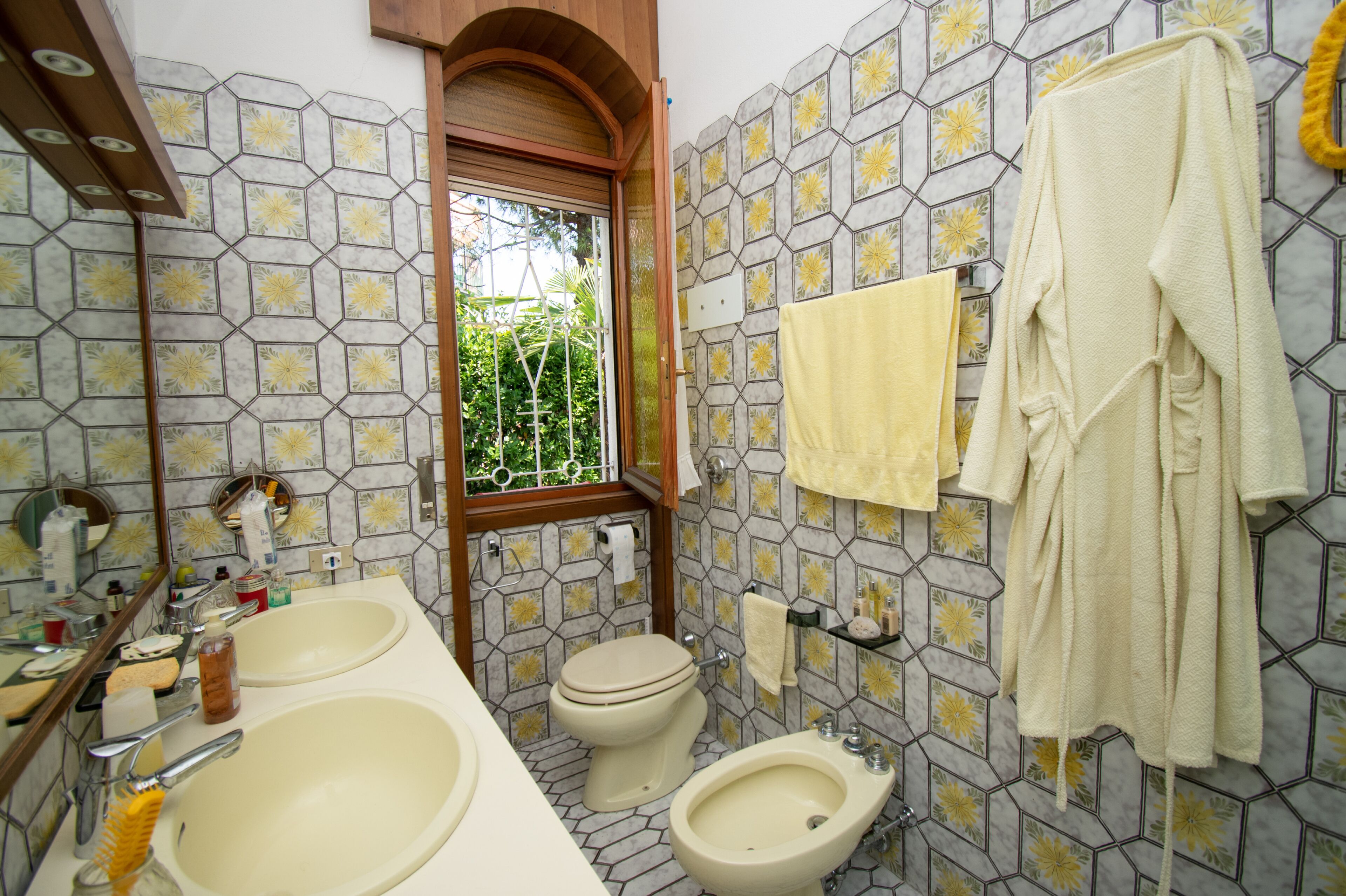 A bathroom with two sinks, a toilet, and a bidet. There is a window with a view of greenery outside.