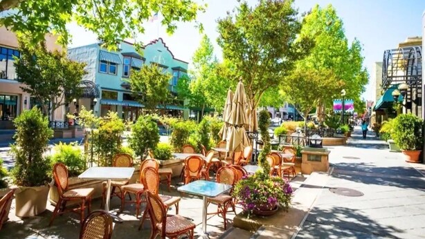 Ein Straßencafé mit Korbstühlen und Tischen, umgeben von Grünflächen und Gebäuden.