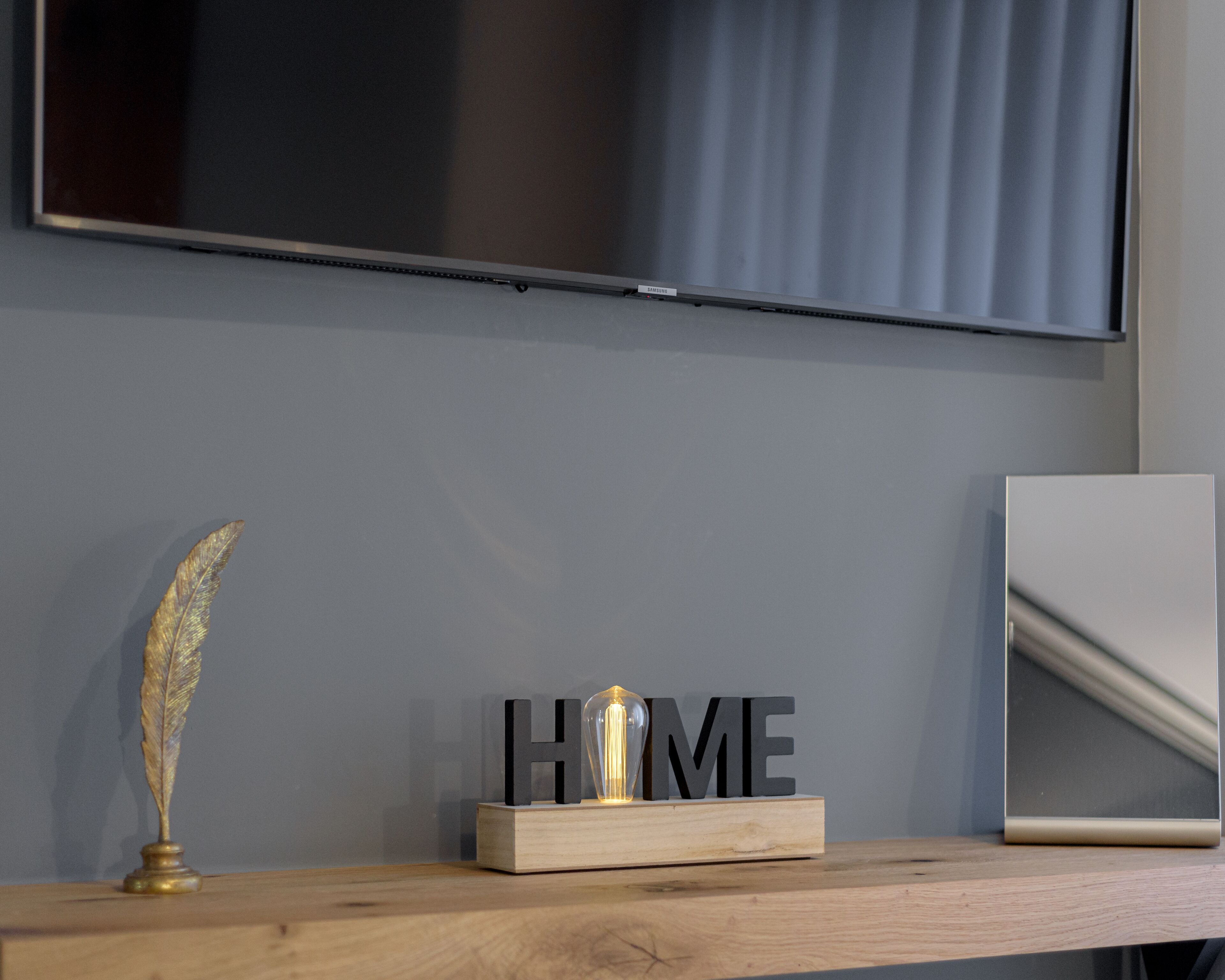 A wooden shelf with a feather quill, a block with the letters "HOME" and a light bulb, and a framed picture leaning against a wall with a television mounted above.