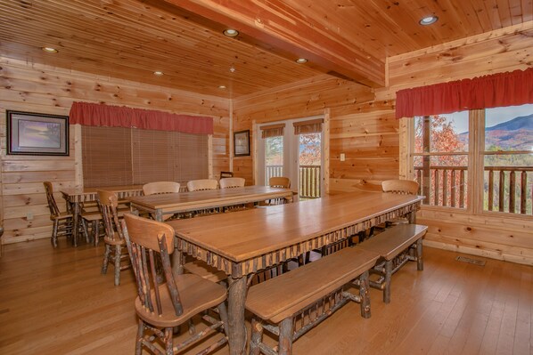 A wooden cabin with a large dining table and benches, framed by wooden walls and windows with red curtains.