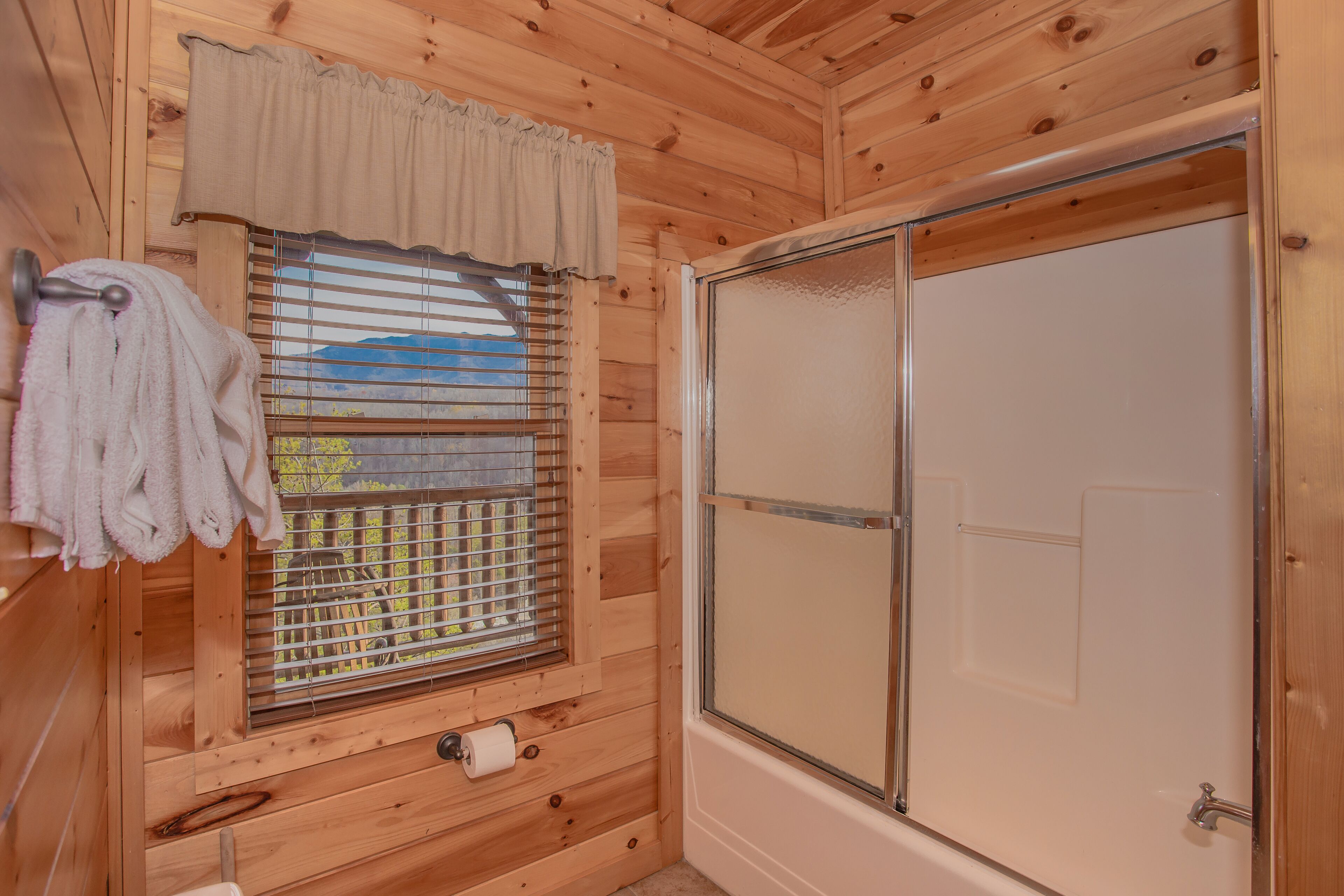 Une salle de bain rustique, avec des murs en bois, une douche, une fenêtre avec des stores et une serviette accrochée à un crochet.
