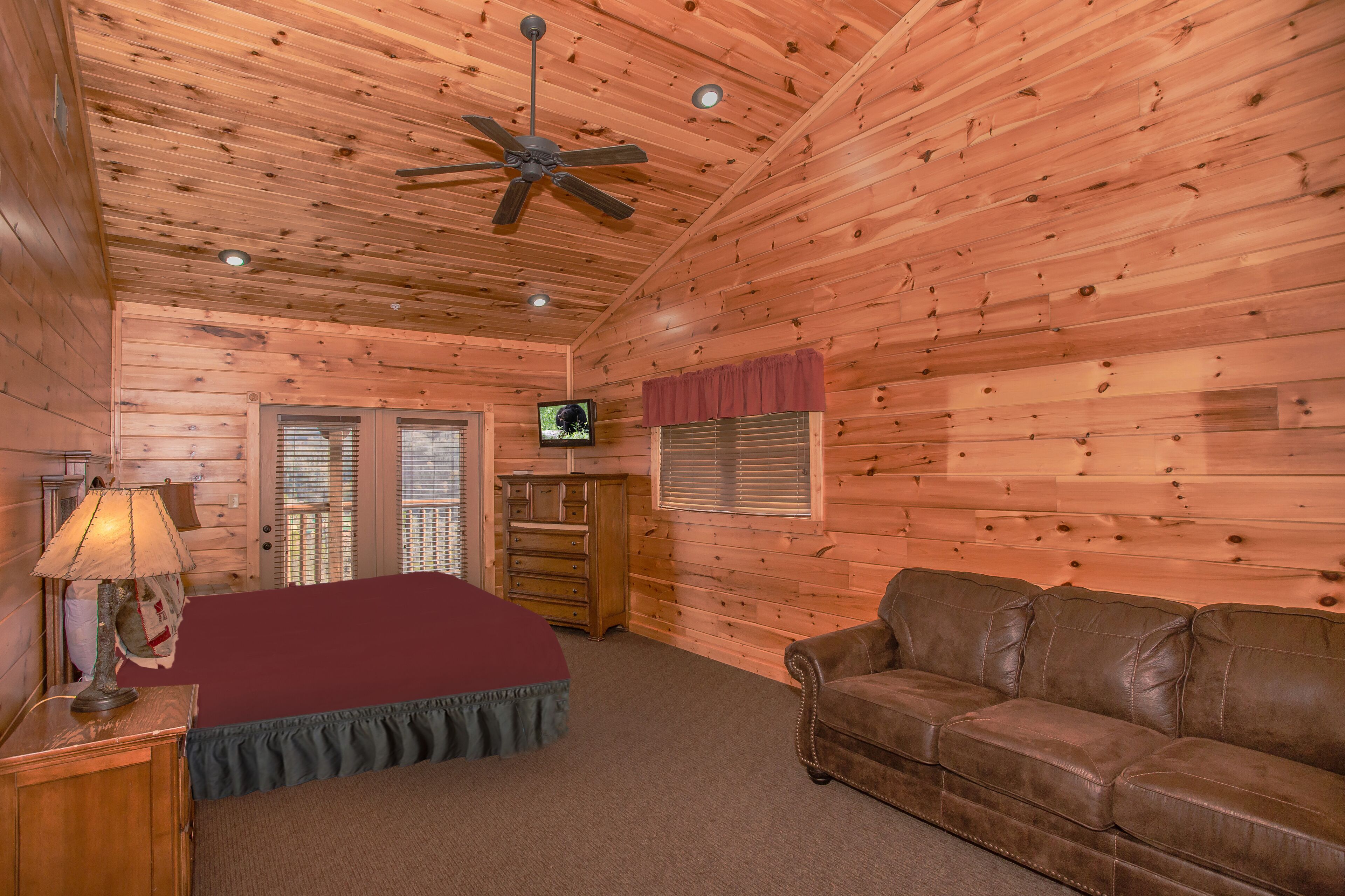 A wooden cabin room with a ceiling fan, a television, a bed with a red blanket, a brown leather couch, and a wooden dresser.