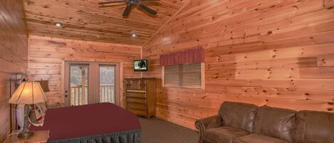 A wooden cabin room with a ceiling fan, a television, a bed with a red blanket, a brown leather couch, and a wooden dresser.
