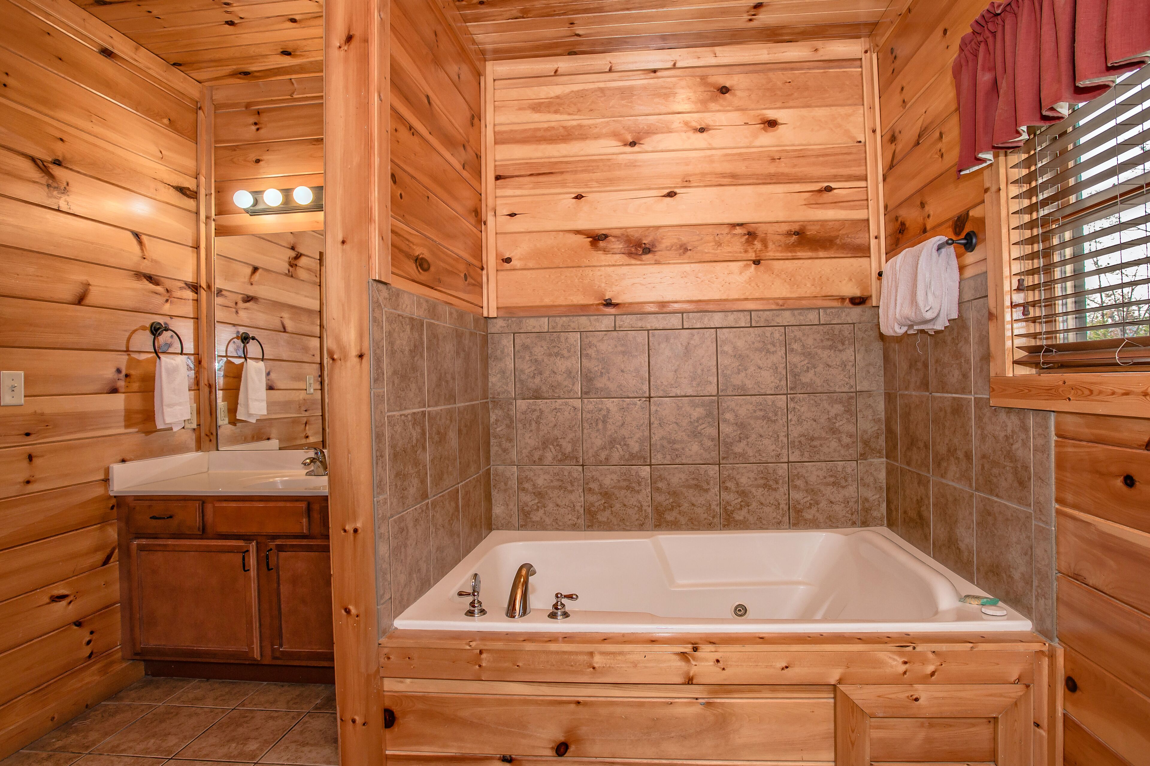 A wooden cabin bathroom with a bathtub, a sink, and a window with blinds.