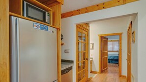 A compact kitchen with a refrigerator, microwave, and dishwasher. A wooden door leads to a bedroom with a bed and a window.