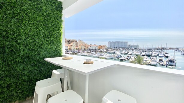 A balcony with a white table and two white stools, a green wall, and a marina view.