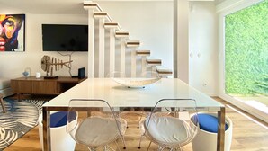A modern living room with a glass dining table, two chairs, a staircase, a flat-screen TV, and a large window with a view of greenery.