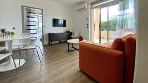 Un salon moderne comprenant un canapé, une table à manger avec des chaises, un téléviseur fixé au mur et une porte-fenêtre coulissante donnant sur un espace extérieur.