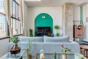A modern living room with a grey sofa, a glass coffee table, and potted plants. - Sosuite | 1BR Penthouse w W/D, Gym, On-Site Bar (Philadelphia)