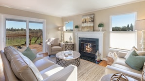 A spacious living room with a fireplace, comfortable seating, and large windows offering a view of the outdoors.