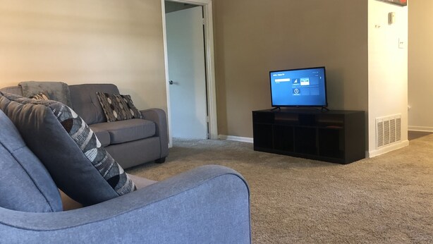 Una sala de estar con un sofá gris, un televisor sobre un mueble y una puerta que da a otra habitación.