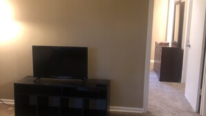 Una habitación con televisión sobre un soporte negro, paredes color beige y piso alfombrado.