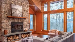 A cozy living room with a stone fireplace, a large window with a view of trees, a comfortable sofa, and a wooden ceiling. - Maine Stay- Contemporary Mountain Home, Spectaular Outdoor Space! (Newry)