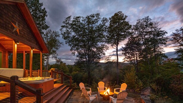 A stone patio with a fire pit and Adirondack chairs, surrounded by trees and a wooden deck with a hot tub. - Maine Stay- Contemporary Mountain Home, Spectaular Outdoor Space! (Newry)