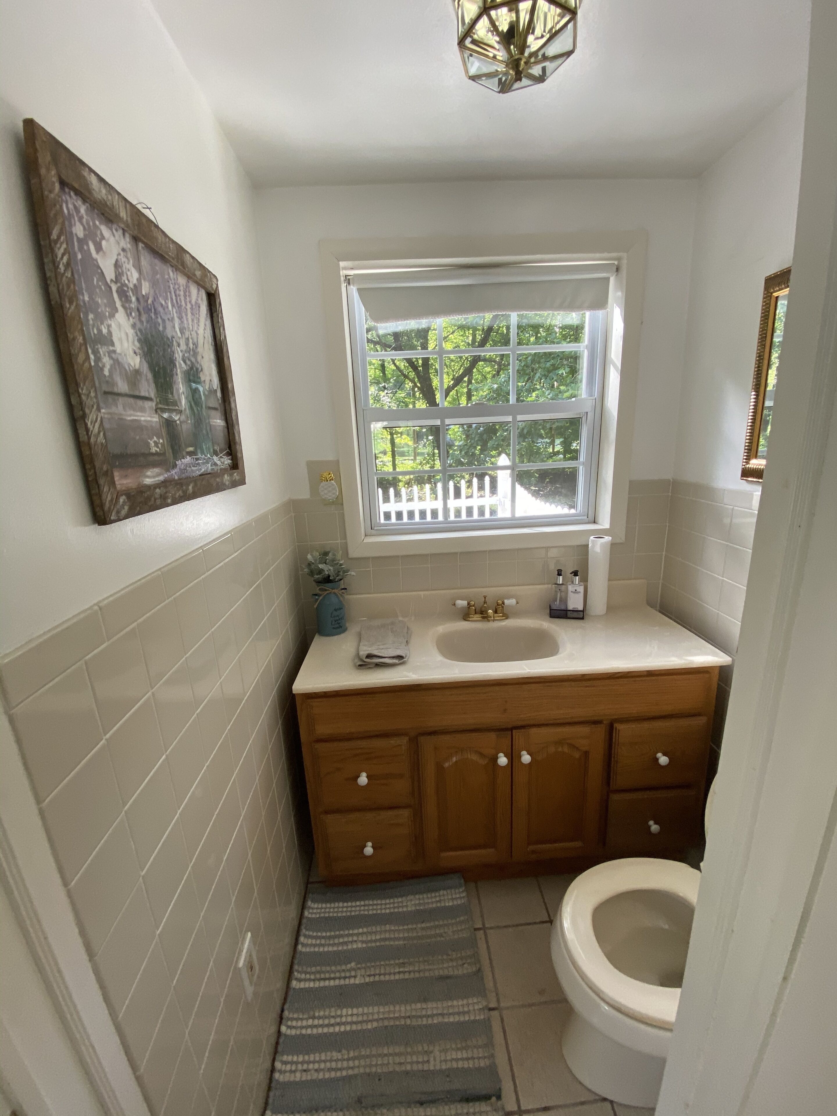 A bathroom with a white sink, wooden vanity, a window with a view of trees, and a single toilet.