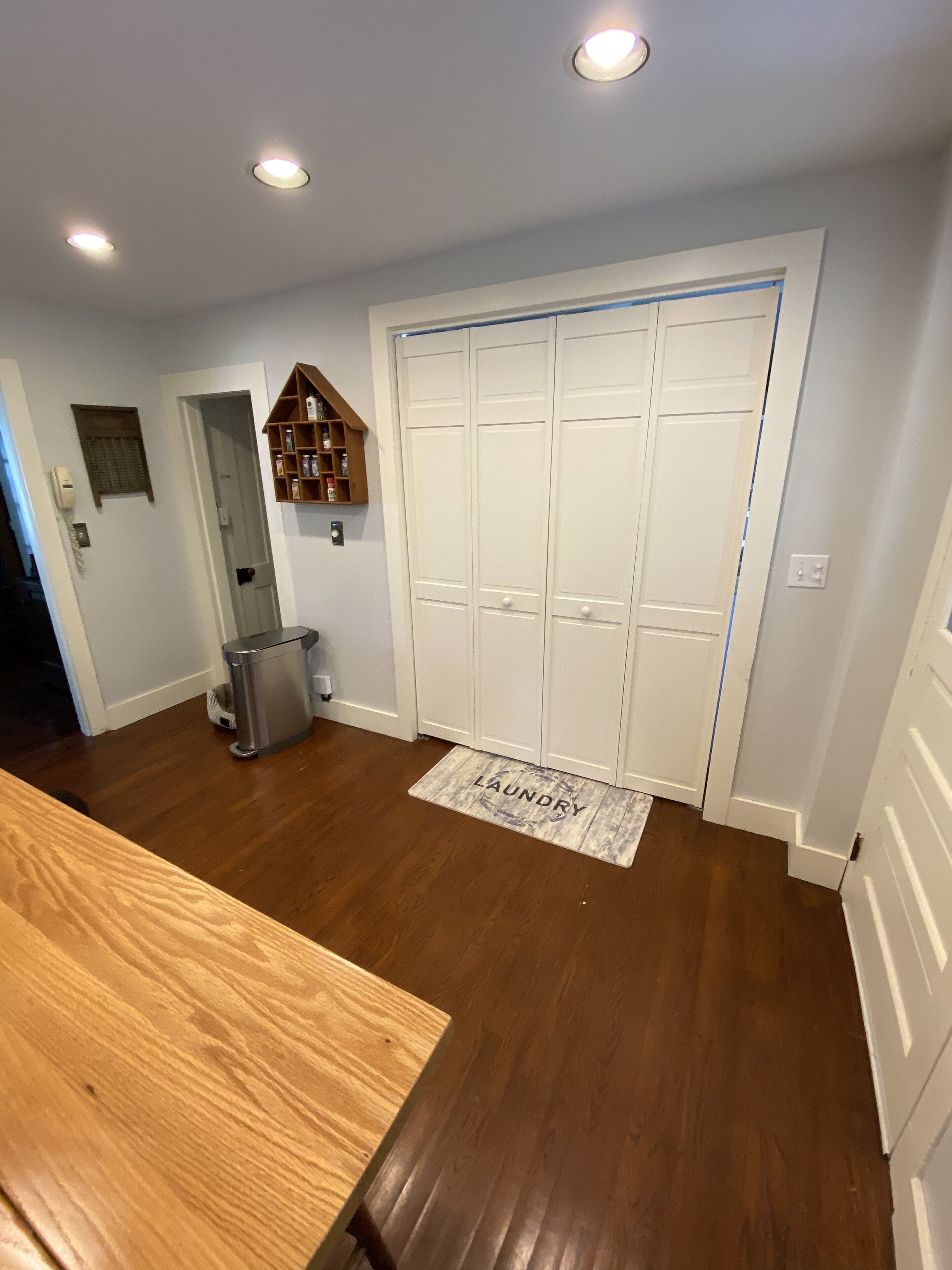 A room with a wooden floor, a white door with a 'laundry' mat, a trash can, and a small wooden shelf with items.