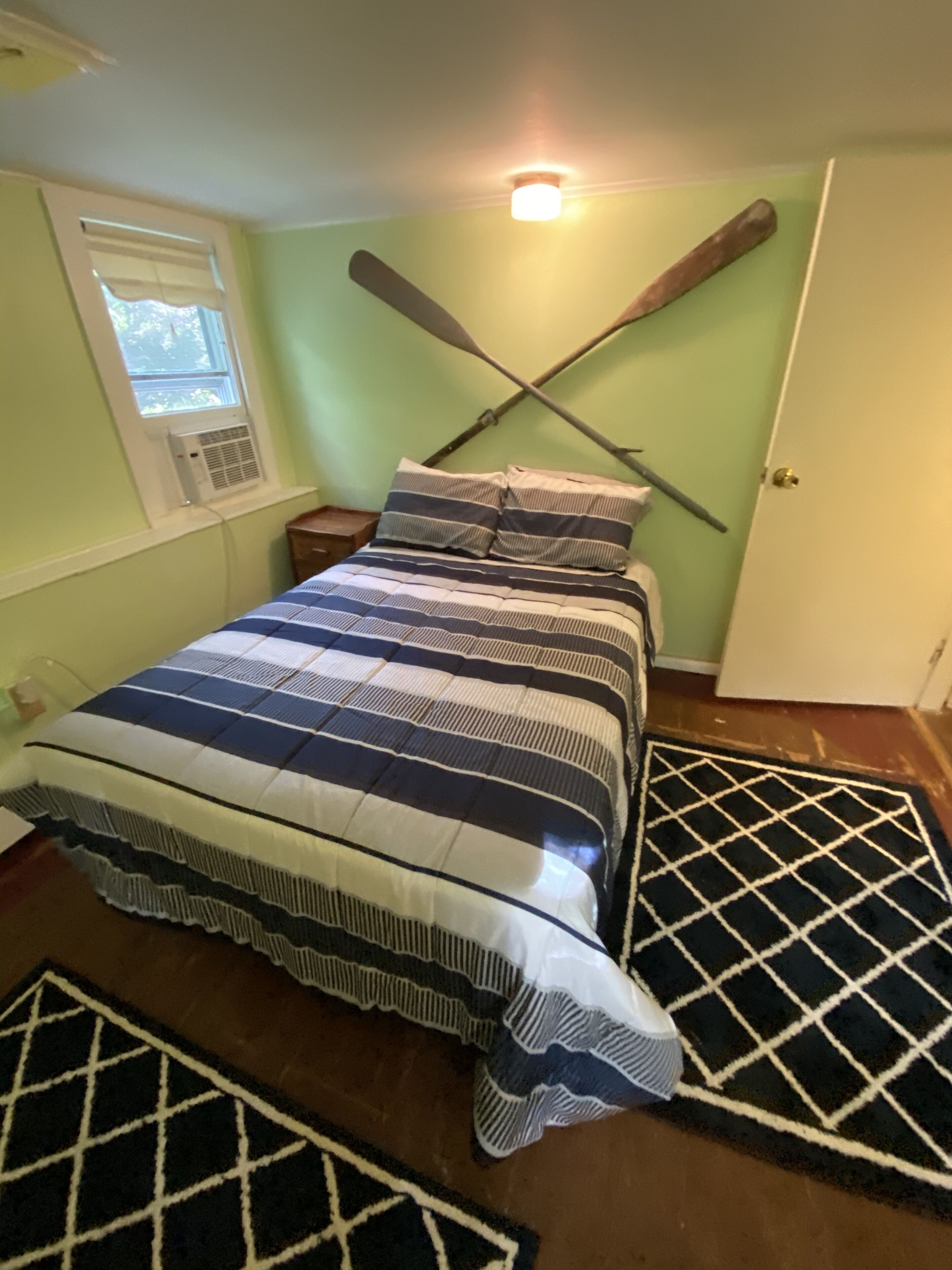 Une chambre à coucher avec un lit, deux oreillers, une fenêtre avec des stores, un ventilateur de plafond et une porte.