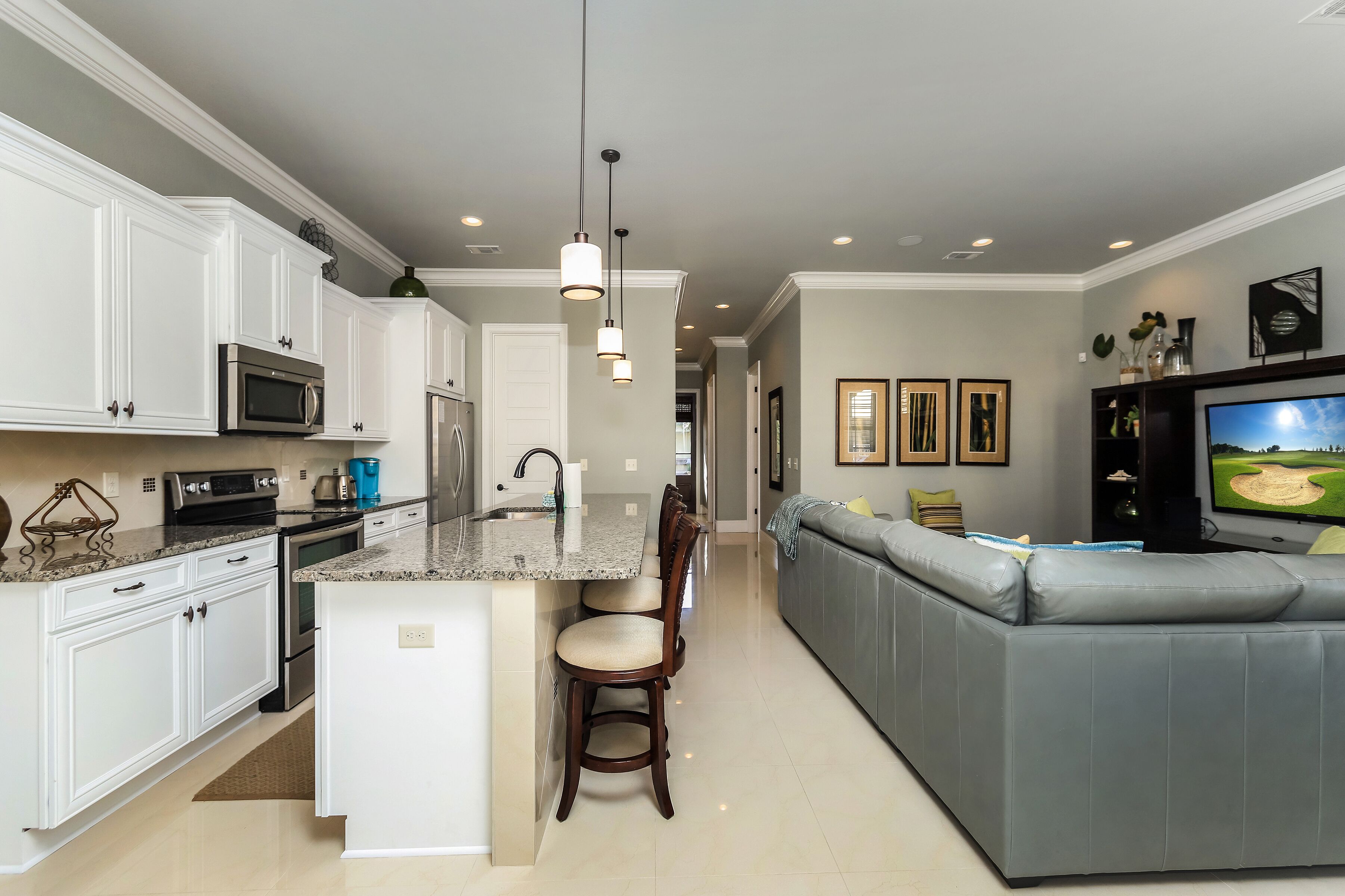 A modern kitchen with white cabinets, a granite countertop, and stainless steel appliances. There is a grey sectional sofa and a flat-screen TV in the living area.