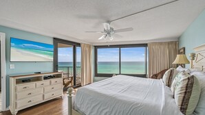 Une chambre dotée d’un grand lit, d’un ventilateur de plafond, d’une commode et offrant une vue sur l’océan grâce à de grandes fenêtres.