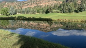 Un paisaje sereno con un lago transparente que refleja árboles y colinas, rodeado de un campo verde exuberante y una montaña al fondo.