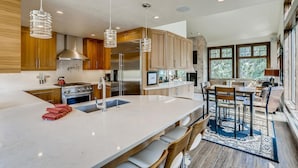 A modern kitchen with wooden cabinets, stainless steel appliances, a large island with bar stools, and a view to the outside. - Wellington Haus: Dog Friendly Home, Hot Tub, Views (Breckenridge)
