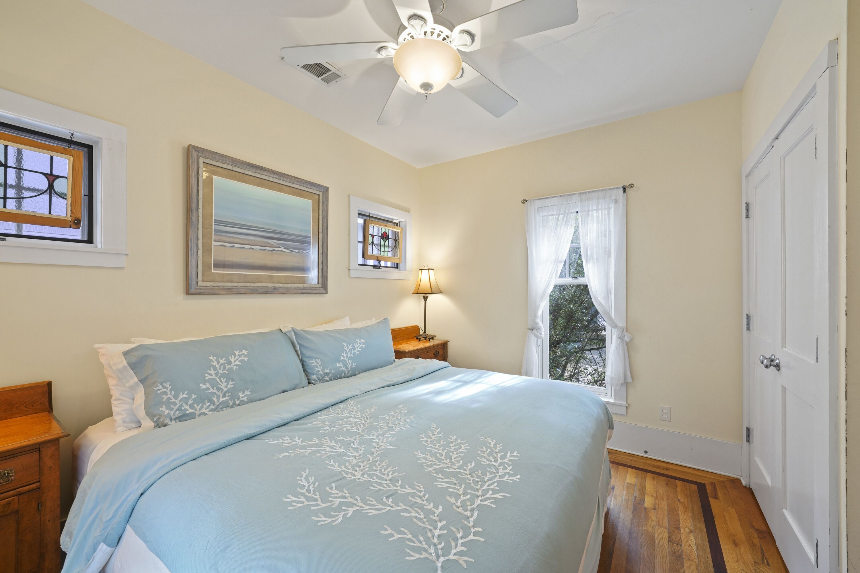 A bedroom with a bed, a ceiling fan, a nightstand, a lamp, a window with curtains, and a door.