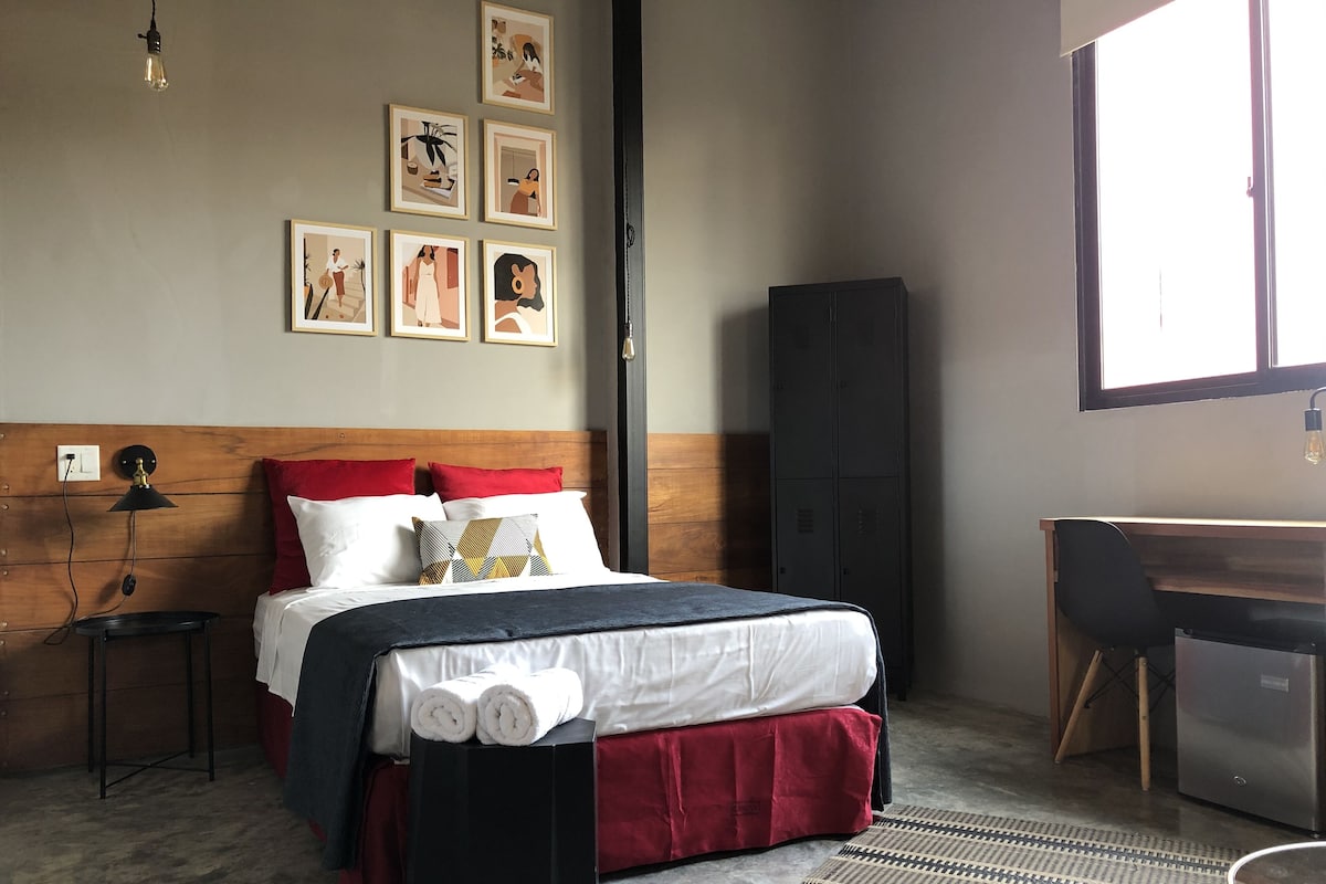A neatly arranged bedroom with a wooden headboard, a bed with white and red bedding, a black speaker, a desk with a chair, and a wardrobe.