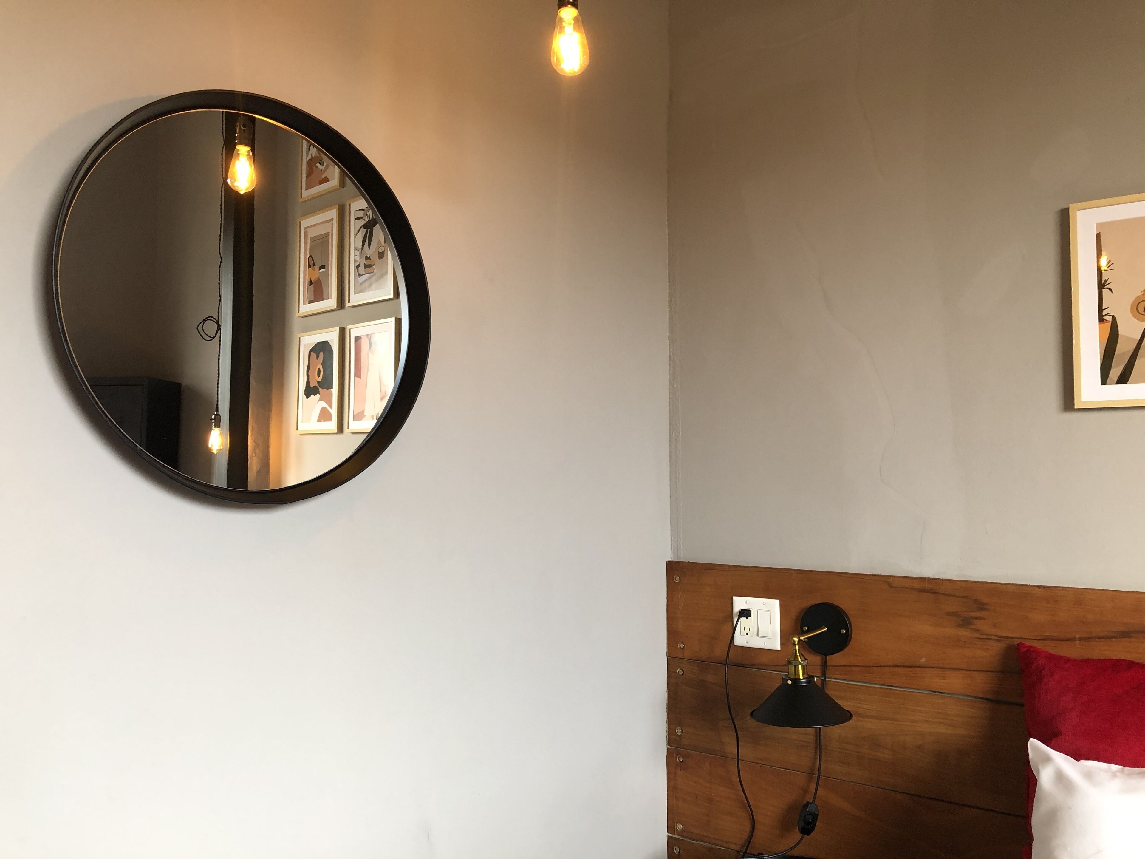 A round mirror on a wall with a light bulb above it, a wooden headboard with a red pillow, and a framed picture.
