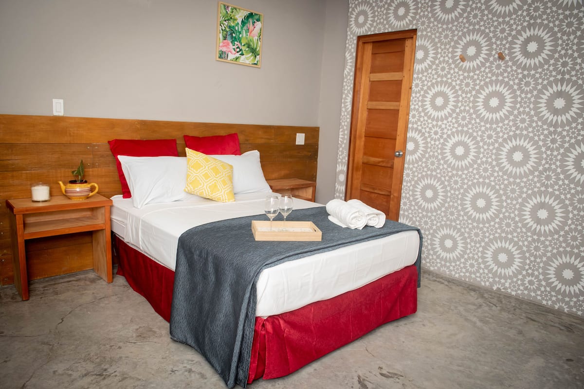 A neatly made bed with a wooden headboard, a bedside table with a plant, and a wall with a geometric pattern.