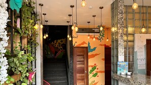 A hallway with a staircase, a wall with colorful murals, hanging light bulbs, and a counter with a sign. - Canova (Panama City)