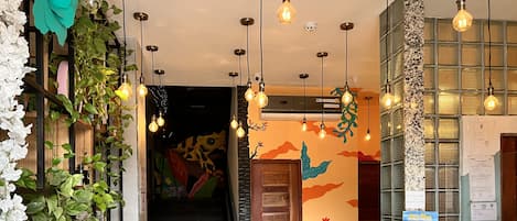 Un pasillo con una escalera, un muro con murales coloridos, focos colgantes y un mostrador con un letrero.