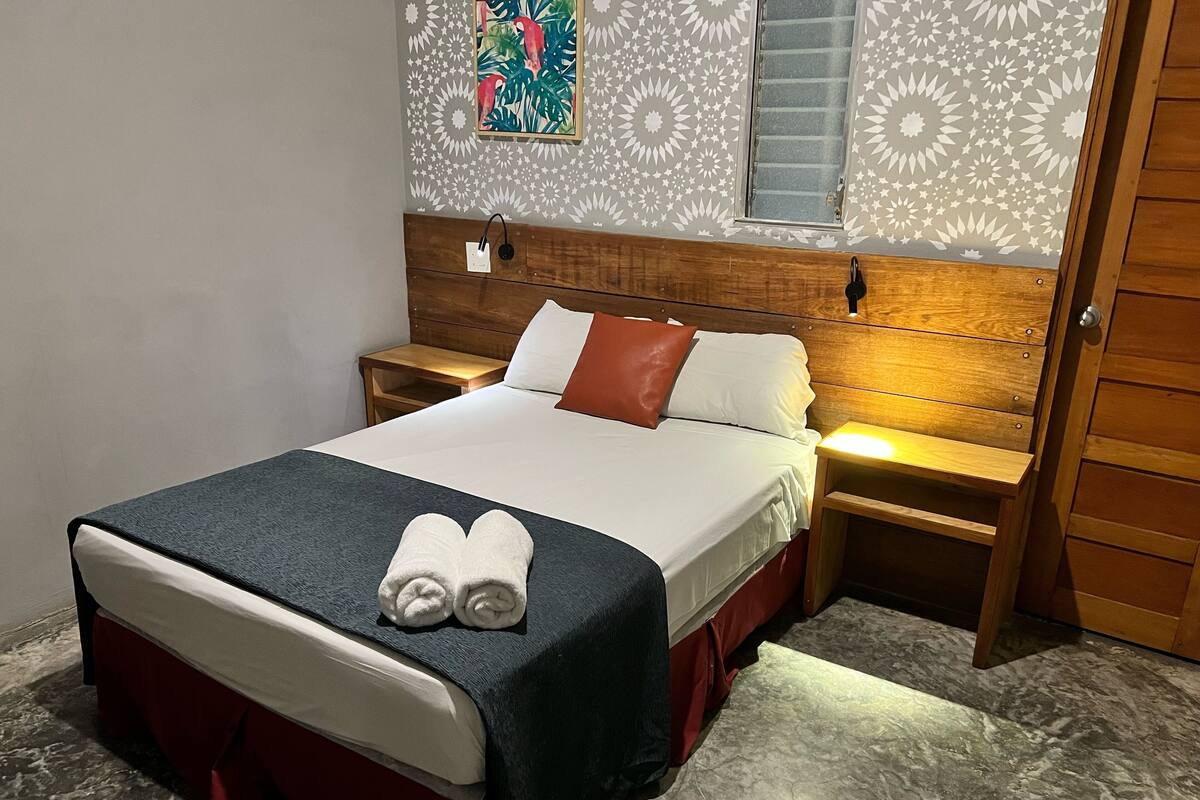 A neatly made bed with a blue blanket, white pillows, and a pair of folded towels. There is a wooden headboard, a nightstand with a lamp, and a wall with a geometric pattern.
