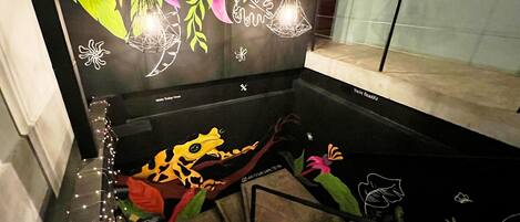 Un interior moderno con una pared negra decorada con diseños de hojas y flores de colores, una mesa de vidrio con una decoración de rana, y una escalera que conduce a un nivel superior.