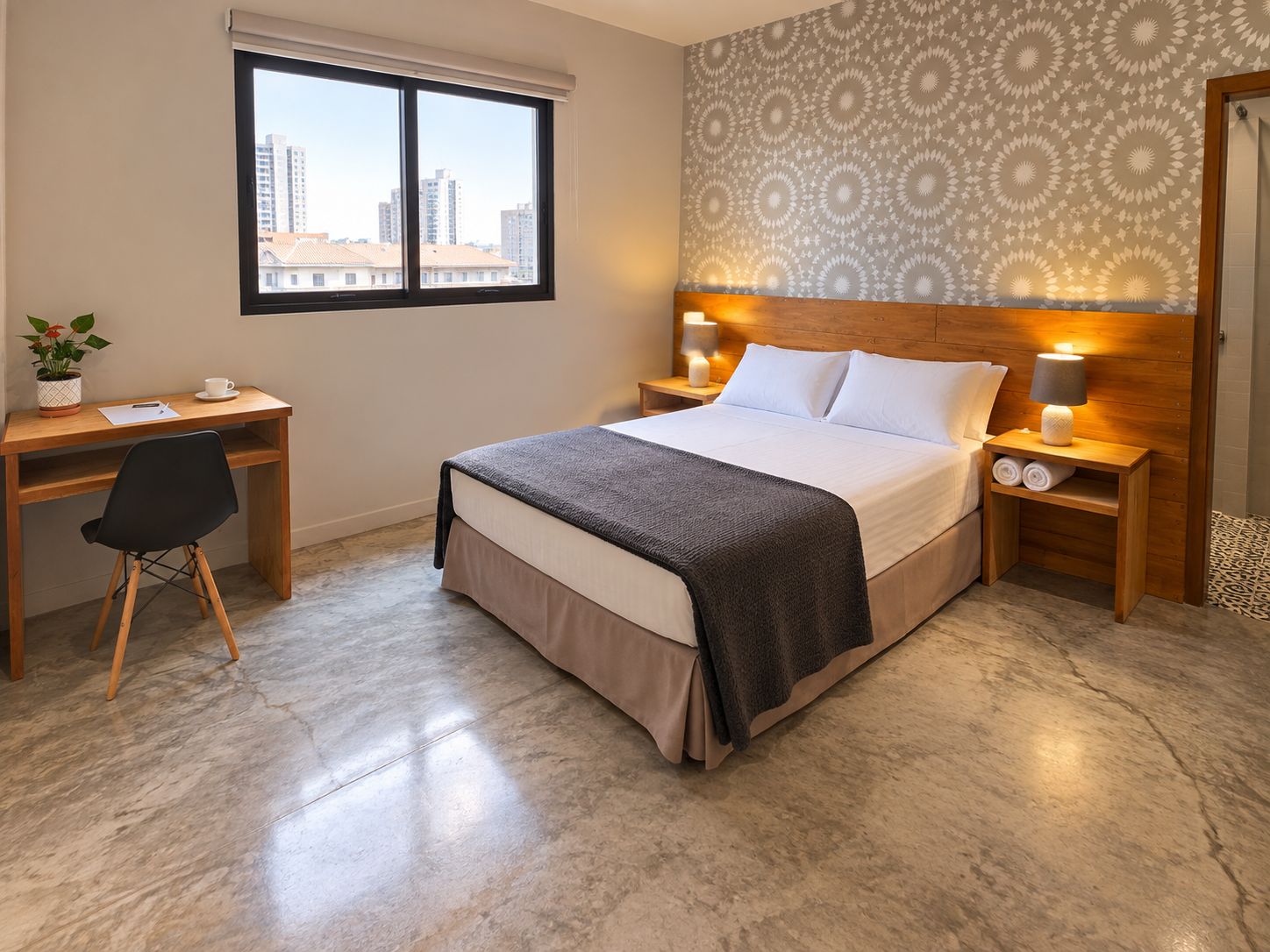 A hotel room with a bed, a desk, a chair, a nightstand with a lamp, and a window with a view of buildings.