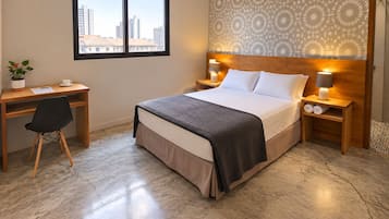Una habitación de hotel con cama, escritorio, silla, mesita de noche con lámpara y ventana con vistas a edificios.