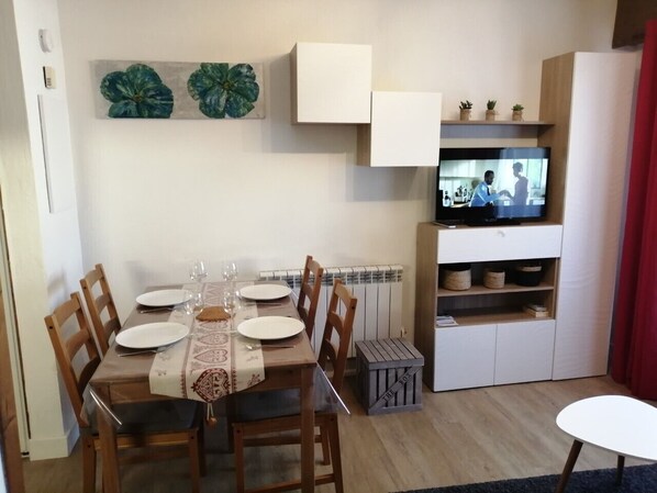 Uma sala de jantar com uma mesa posta para quatro pessoas, uma televisão e um móvel com prateleiras.
