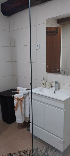 Un baño con un lavamanos blanco, espejo y paredes cubiertas de azulejos. Hay un bote de basura negro y un perchero con una toalla y un lazo rosa.