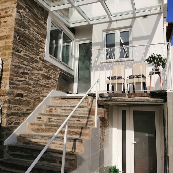 Una casa moderna con fachada de piedra, un invernadero de cristal y una escalera blanca que lleva a un balcón con mobiliario exterior.