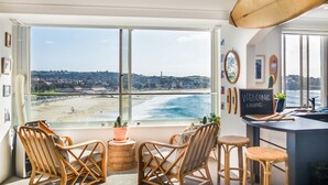 Una stanza con una grande finestra vista spiaggia, sedie in vimini, un piccolo angolo bar e un'asse da surf al soffitto.