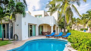 Una piscina con agua azul, rodeada de palmeras y un edificio blanco con un patio.