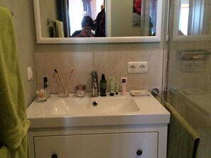 A bathroom with a white sink, a mirror above it, and toiletries on the counter. There is a green towel on the left and a shower area to the right.