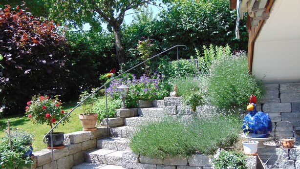 A stone staircase leading to a garden area with potted plants and a decorative rooster statue. - Ferienwohnung, 55 qm Obergeschoss, 1 Separates Schlafzimmer (Bad Endorf)