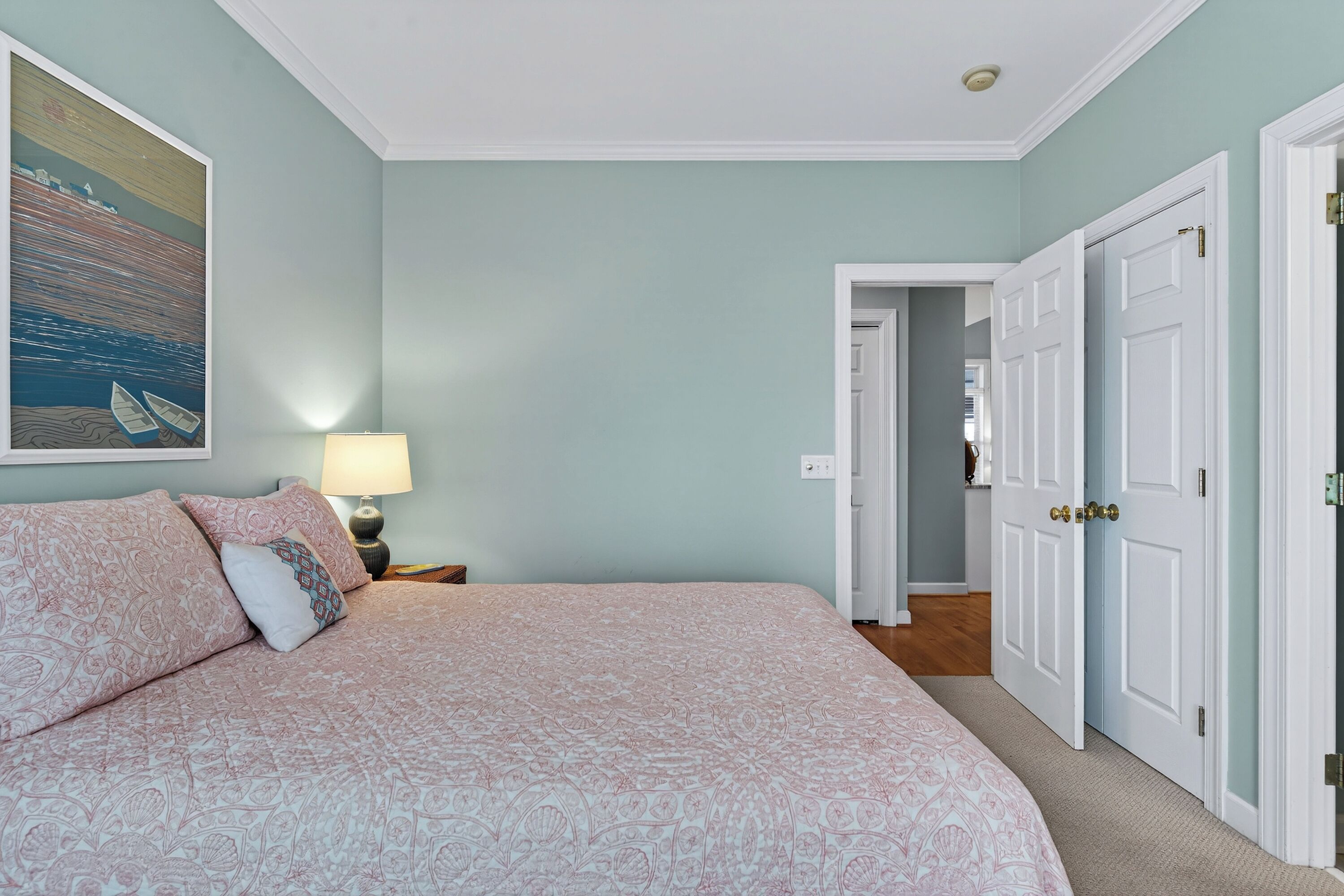 Een slaapkamer met een groot bed, een nachtkastje met een lamp, een schilderij aan de muur en een deur naar een andere kamer.